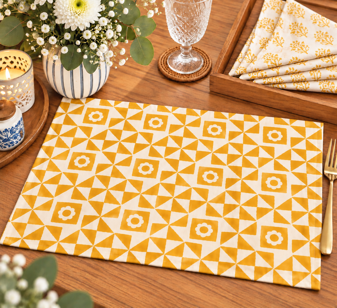 A rectangular placemat with a yellow and white geometric triangle pattern is displayed on a wooden table, surrounded by dining props including a glass, candle, flowers, and folded napkins.