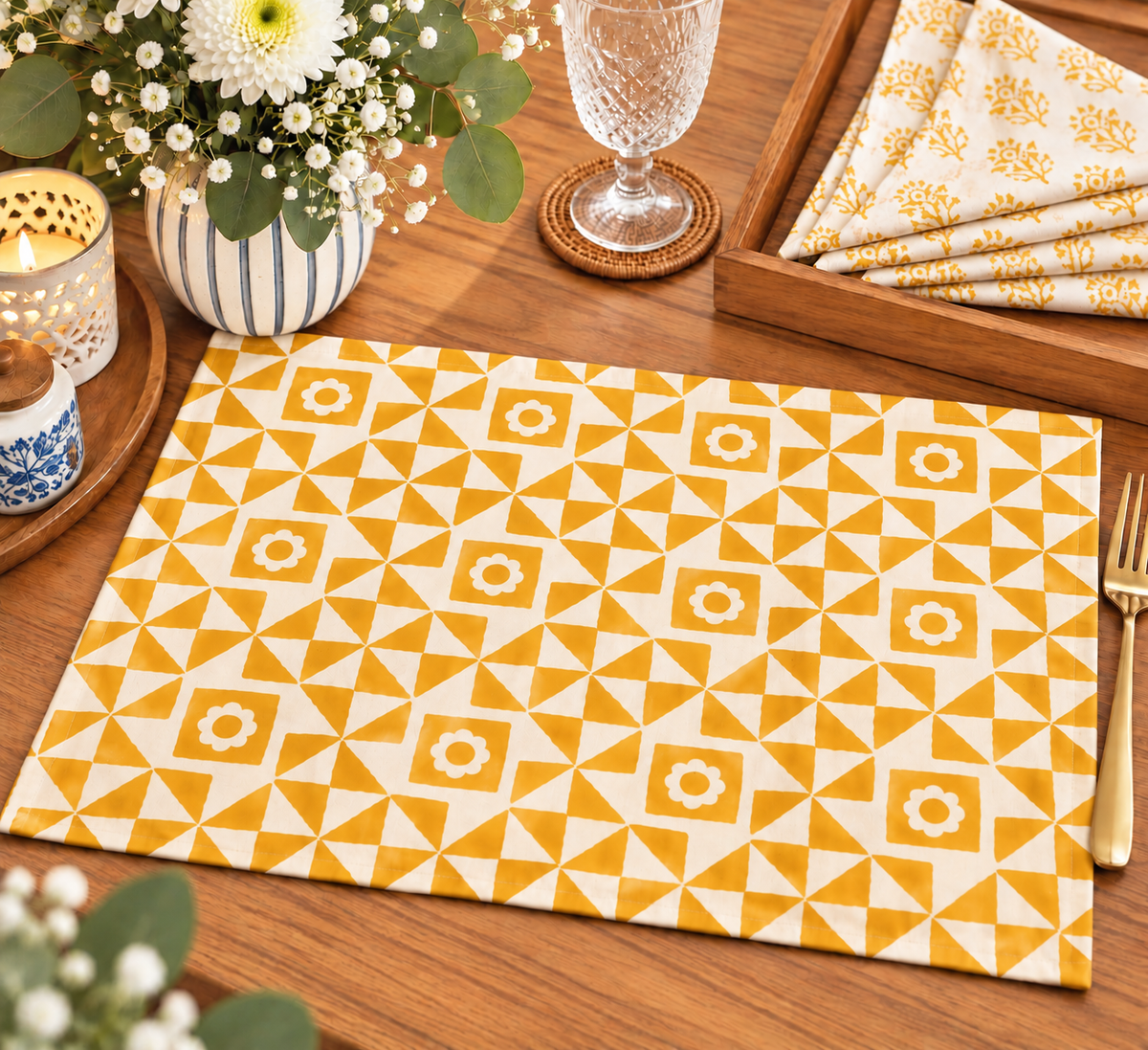 A rectangular placemat with a yellow and white geometric triangle pattern is displayed on a wooden table, surrounded by dining props including a glass, candle, flowers, and folded napkins.