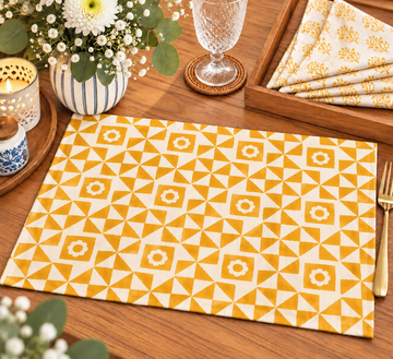 A rectangular placemat with a yellow and white geometric triangle pattern is displayed on a wooden table, surrounded by dining props including a glass, candle, flowers, and folded napkins.