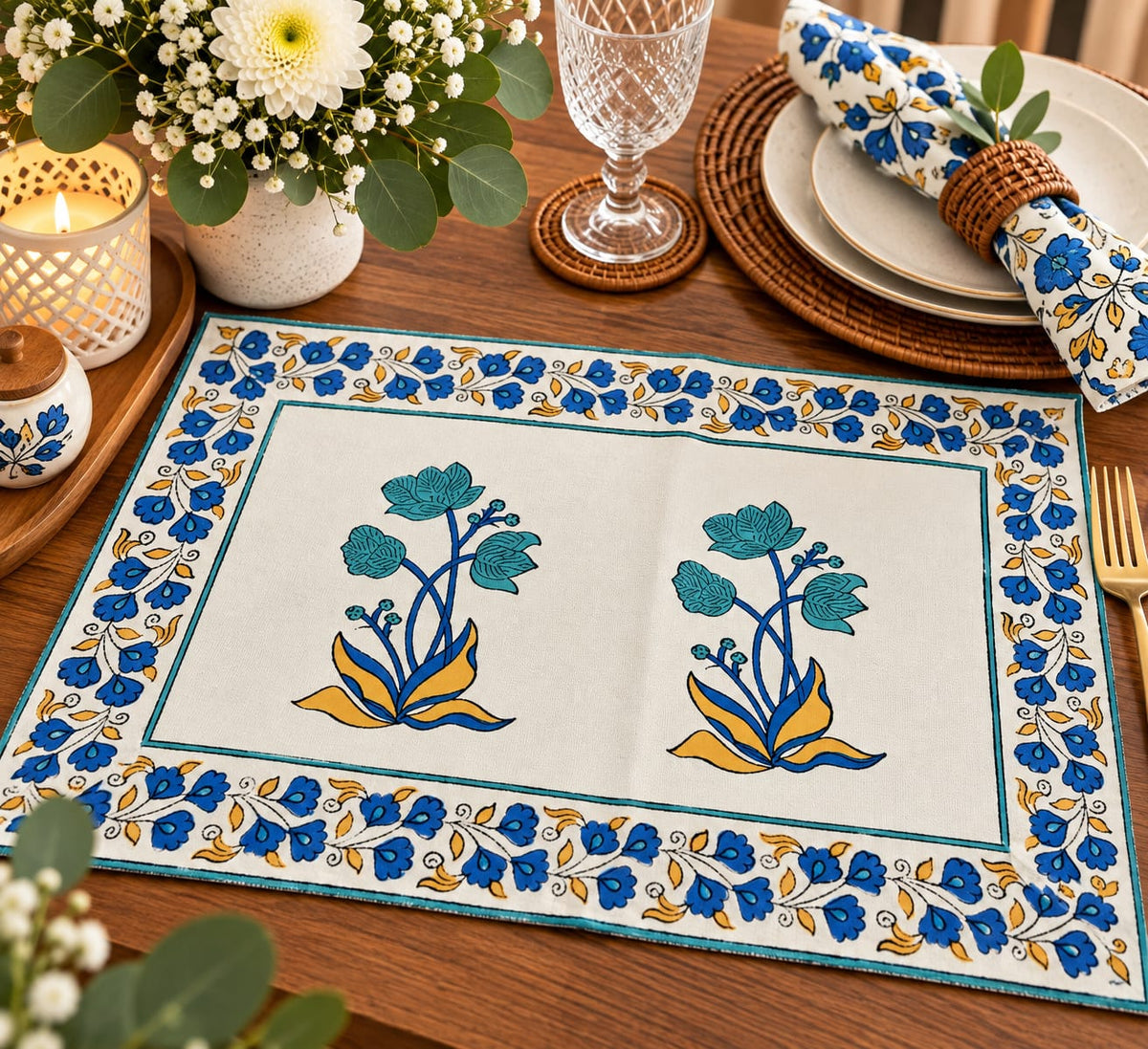 A rectangular embroidered table placemat with a cream background, blue floral border, and two matching floral motifs in the center, styled on a wooden dining table with plates, a glass, and a candle.