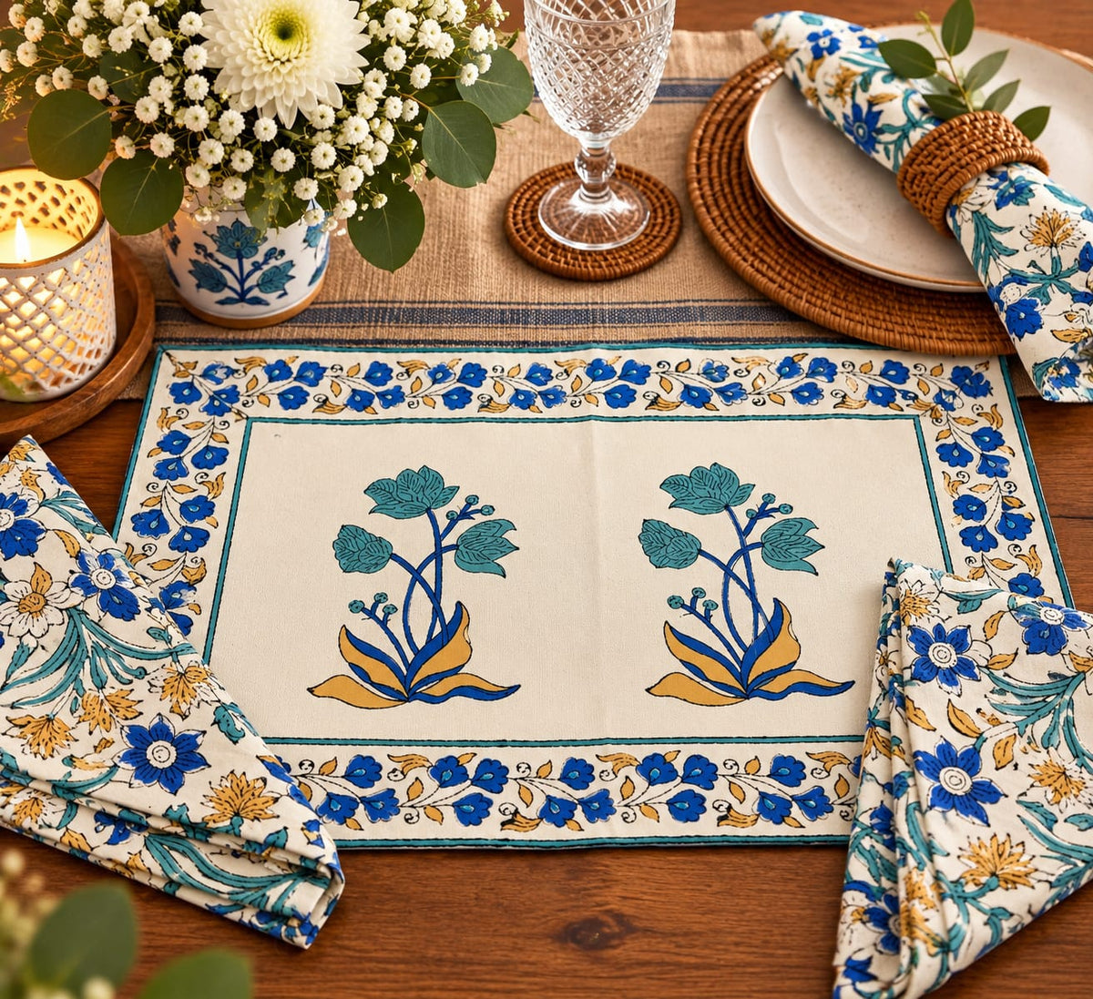A styled table setting featuring a rectangular cream placemat with blue and mustard floral motifs and matching folded napkins, arranged on a wooden table with a candle, flowers, plates, and a glass.