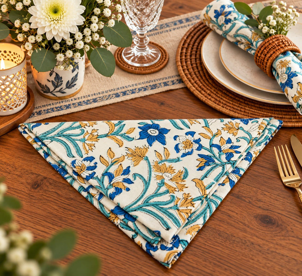 A folded floral napkin with a cream background, blue flowers, and teal leaves is styled on a wooden dining table alongside plates, a glass, flowers, and tableware.