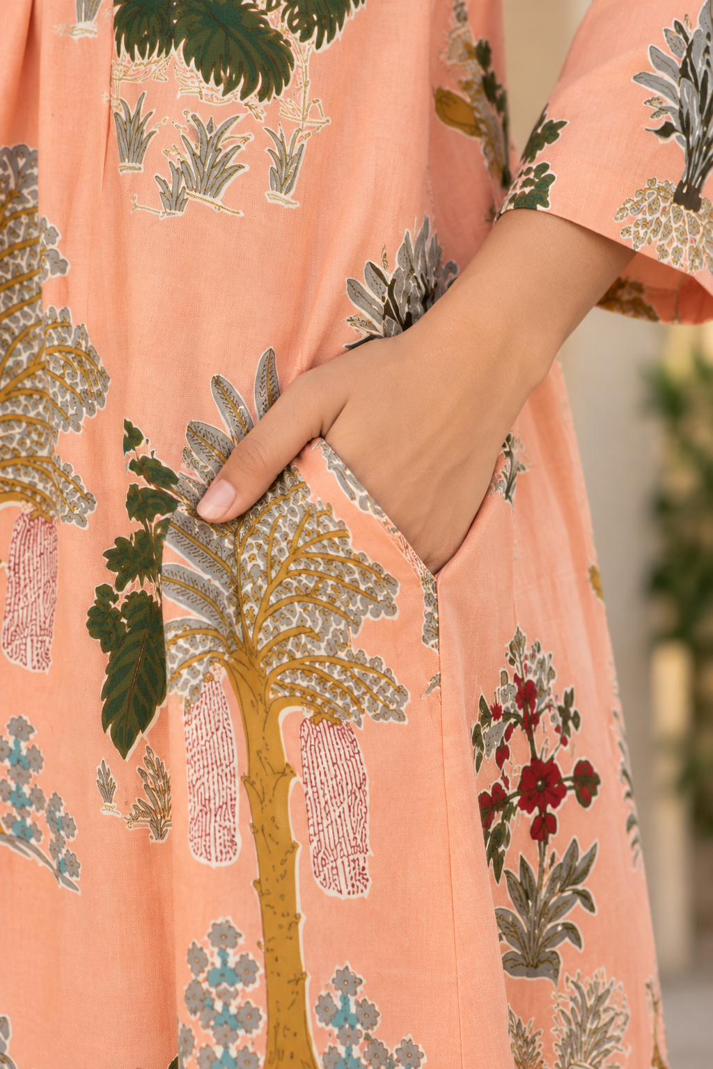 A close-up of a peach-colored dress with a botanical block-print pattern, featuring a hand tucked into a side pocket.