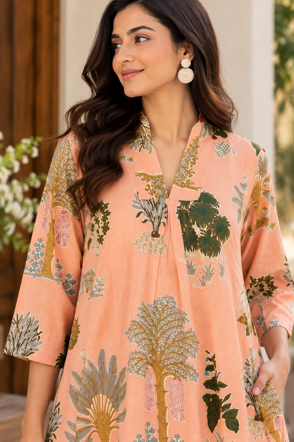 A woman wearing a peach-colored printed kurta with green, gold, and gray botanical patterns, shown in a lifestyle setting with a blurred background.
