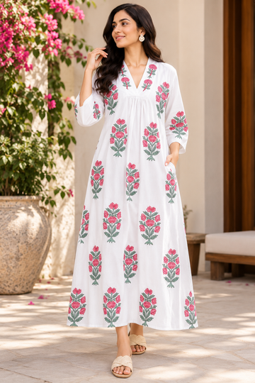 A woman wearing a white maxi dress with pink and green floral embroidery, standing in a sunlit outdoor patio setting with a potted plant in the background.