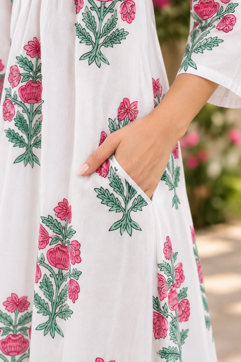 A close-up of a white dress with pink floral and green leaf patterns, featuring a side pocket with a hand inserted, set against a soft-focus outdoor background.