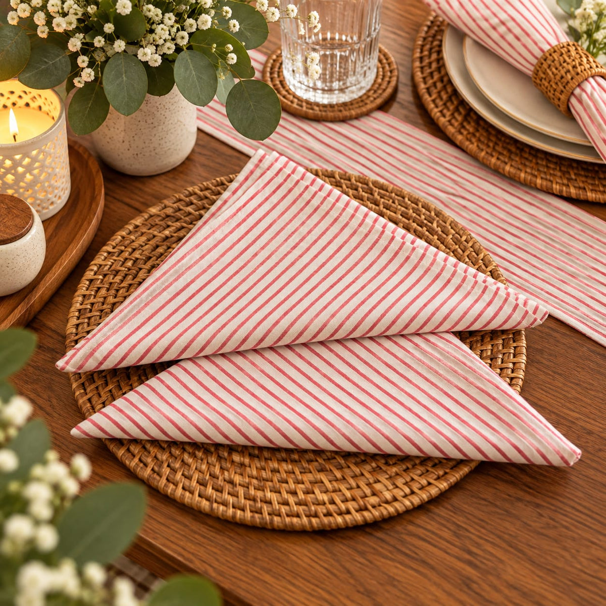 A folded striped cloth napkin in pink and white is arranged on a woven round placemat over a wooden dining table, with candles, flowers, plates, and a glass visible in the setting.