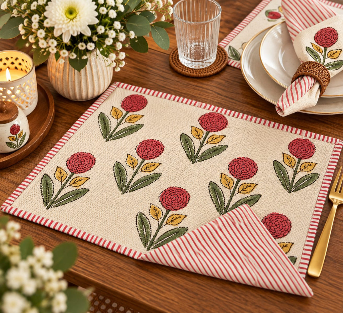 A styled tabletop setting featuring a cream-colored woven placemat with a repeating red and green floral pattern and red-and-white striped borders. A matching folded napkin with red-and-white stripes and a floral motif is placed on the table, alongside plates, a glass tumbler, flowers in a vase, and a lit candle.