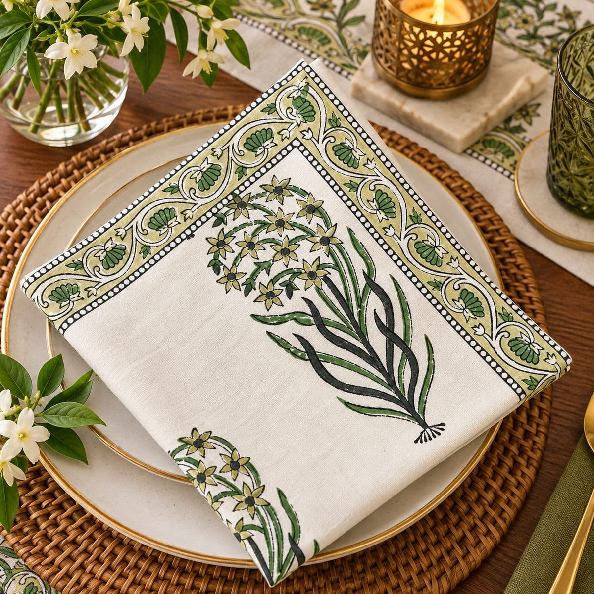 A folded white linen napkin with green botanical and scroll border embroidery, styled on a layered place setting with plates, a woven placemat, flowers, and candle holders.