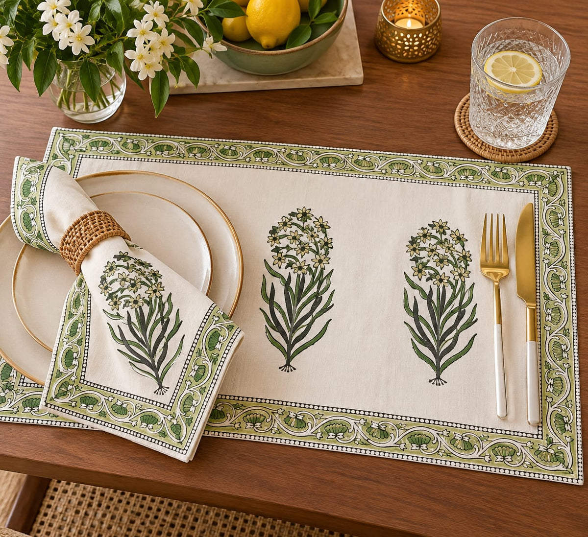 A styled table setting featuring a rectangular cream linen placemat with green botanical floral prints and a matching folded napkin, arranged on a wooden dining table alongside white plates, gold-toned flatware, and decorative flowers and a lemon glass.