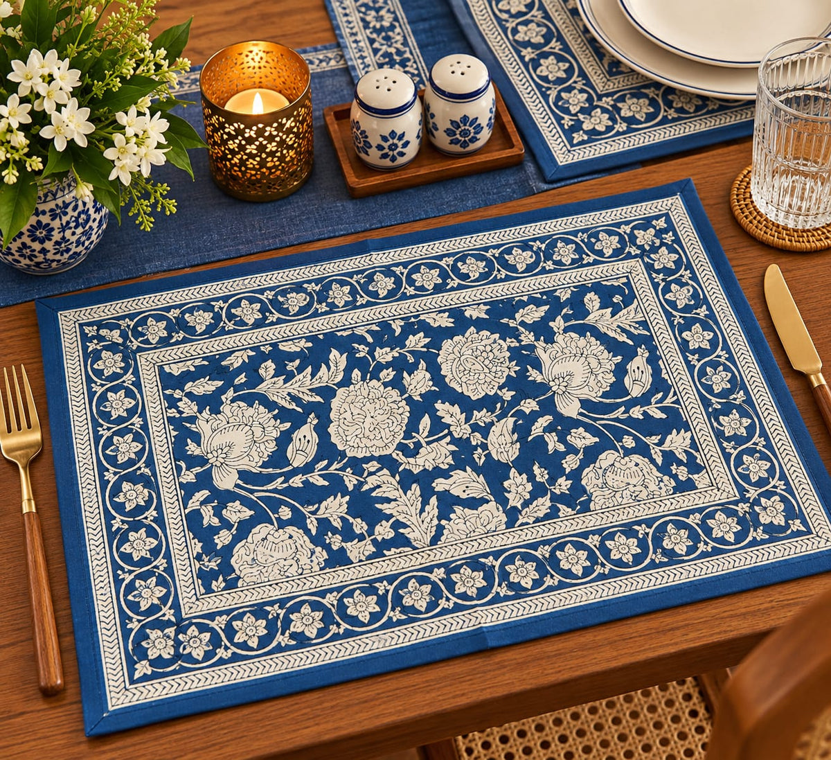 A rectangular blue table placemat with an intricate white floral and vine border pattern, styled on a wooden dining table alongside a glass, salt and pepper shakers, a candle, and a vase of white flowers.