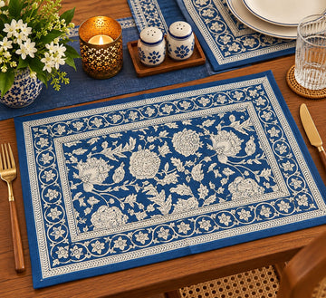 A rectangular blue table placemat with an intricate white floral and vine border pattern, styled on a wooden dining table alongside a glass, salt and pepper shakers, a candle, and a vase of white flowers.
