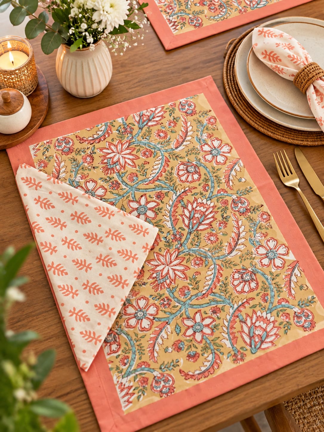 A styled tabletop setting featuring a rectangular place mat with a coral border and a cream background decorated with a detailed floral and vine pattern in coral, teal, and rust. A matching folded napkin with a simple coral leaf print is draped over the corner of the mat.