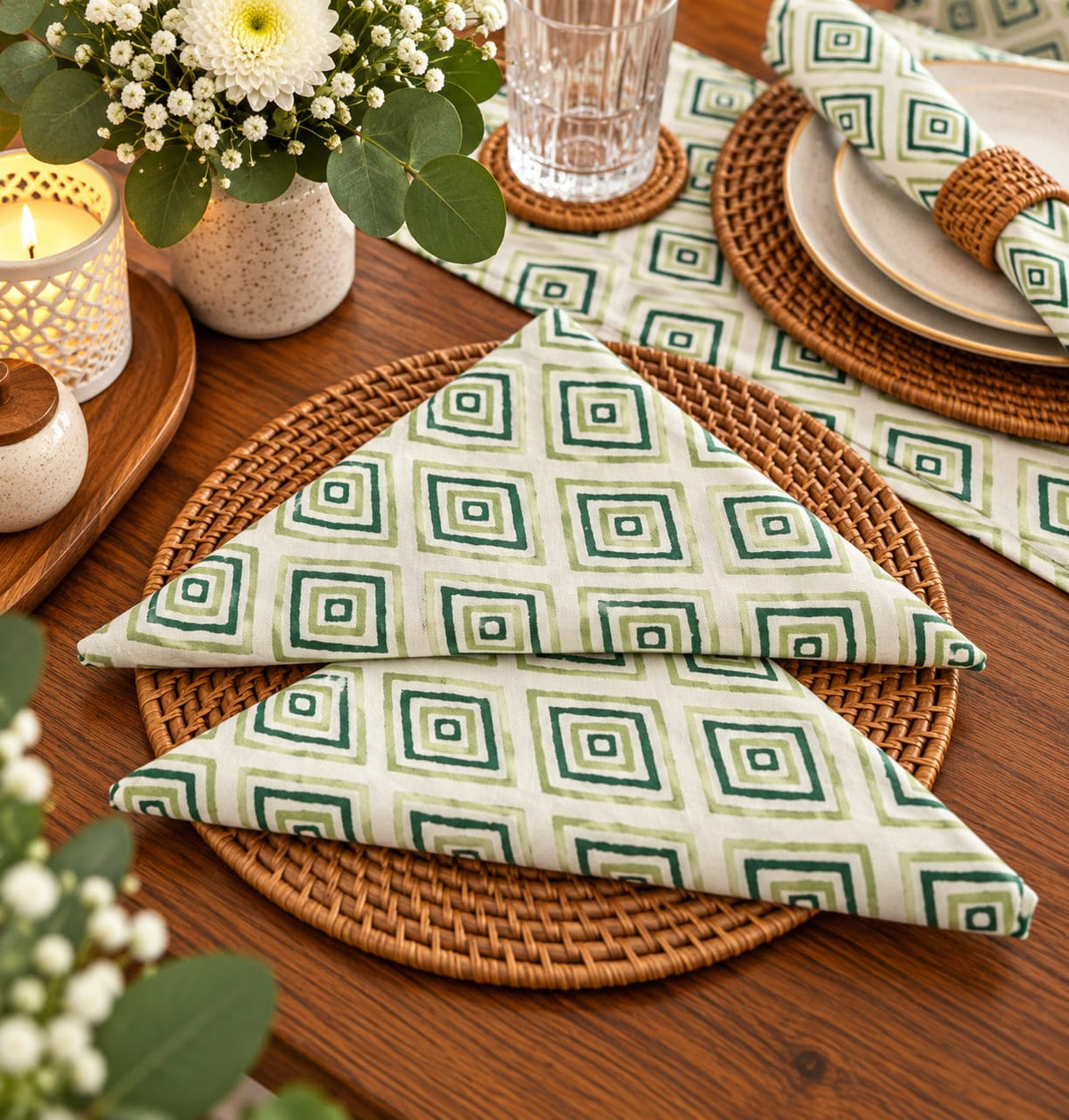 A styled dining table setting featuring a folded cloth napkin with a green geometric square pattern on a cream background, layered over a round woven brown placemat. The scene includes stacked plates, a glass tumbler, a lit candle, and a vase of white flowers.