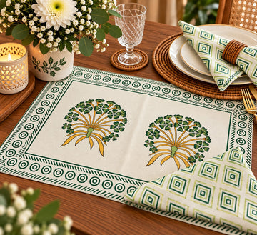 A rectangular table runner laid on a wooden dining table, featuring two green and yellow floral motifs in the center and a decorative border. A matching folded napkin with a geometric green and cream pattern is placed beside it, along with plates, a glass, flowers, and candlelight in a styled dining setting.