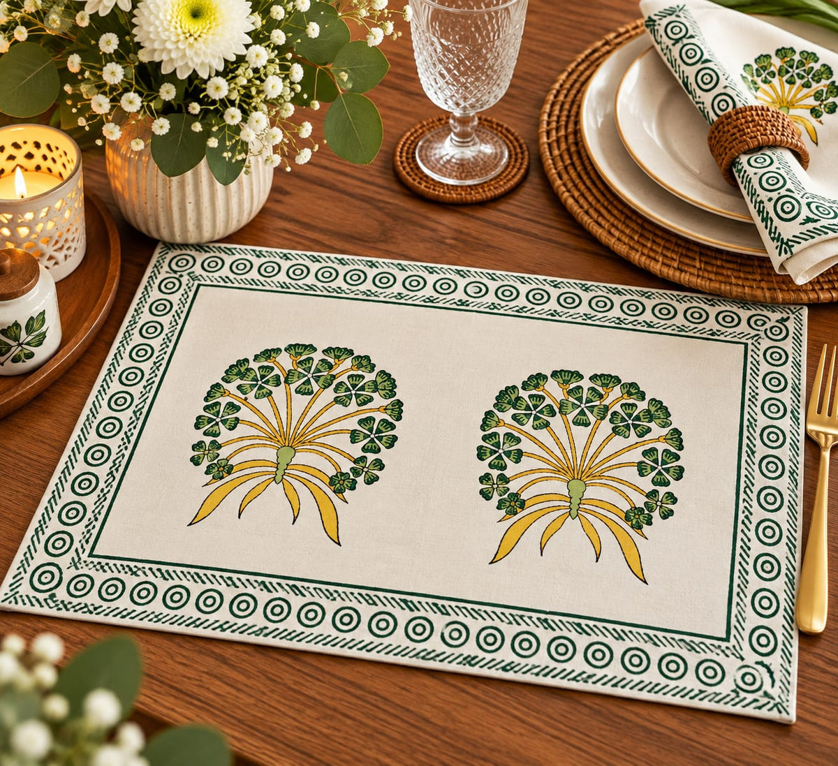 A rectangular table placemat with a cream background, green and yellow floral motifs, and a decorative border, styled on a wooden dining table with plates, a folded matching napkin, flowers, and glassware.