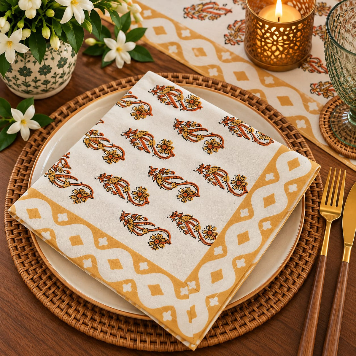 A folded cream-colored cotton napkin with a mustard yellow geometric border and orange-brown floral motifs, presented on a woven placemat over a beige plate on a wooden table.