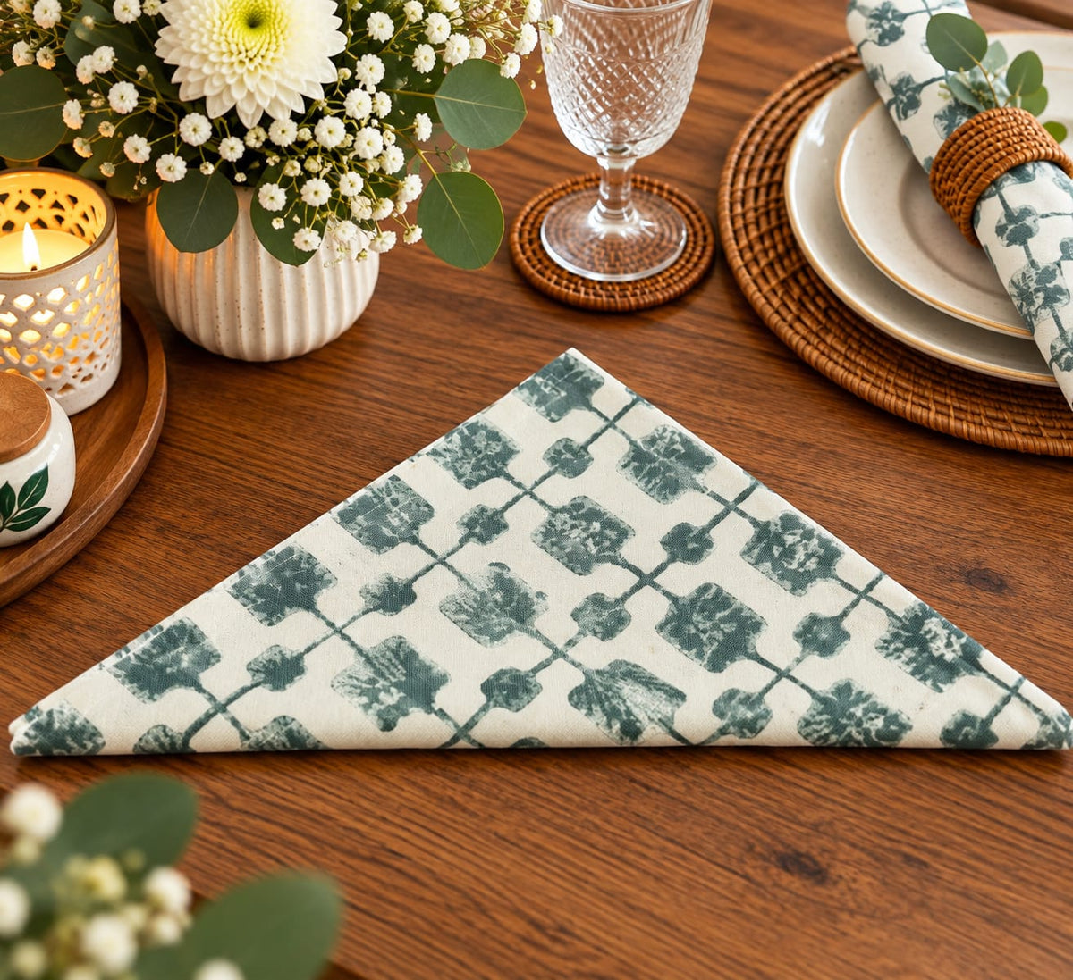A folded cloth napkin with a green and white geometric lattice pattern is displayed on a wooden table, styled with a glass goblet, plates, a candle, and a flower vase.