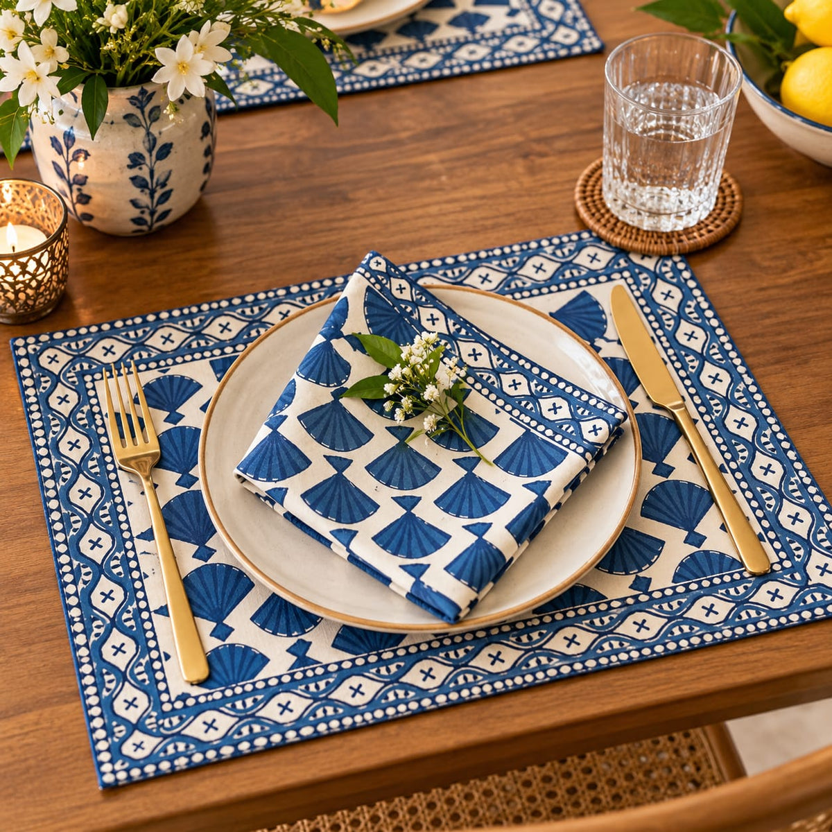A styled dining table setting featuring blue and white patterned placemats, matching napkins, a white plate with gold trim, gold-toned cutlery, and decorative floral accents on a wooden table.