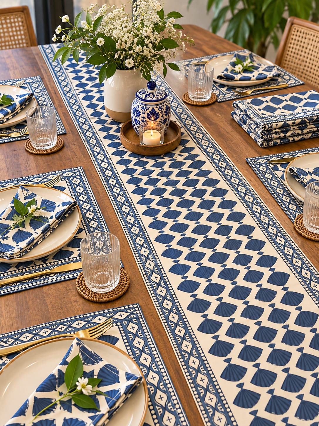 A styled dining table set with a long cream table runner and matching blue patterned placemats, plates, folded napkins, glassware, and a ceramic lidded container with a candle. The scene is photographed in a real dining room environment with wooden chairs and plants visible.
