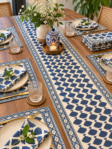 A styled dining table set with a long cream table runner and matching blue patterned placemats, plates, folded napkins, glassware, and a ceramic lidded container with a candle. The scene is photographed in a real dining room environment with wooden chairs and plants visible.