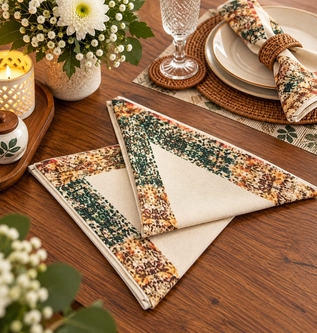 Two folded patterned cloth napkins on a wooden table, styled with plates, a glass, flowers, and woven placemats in a warm dining setting.