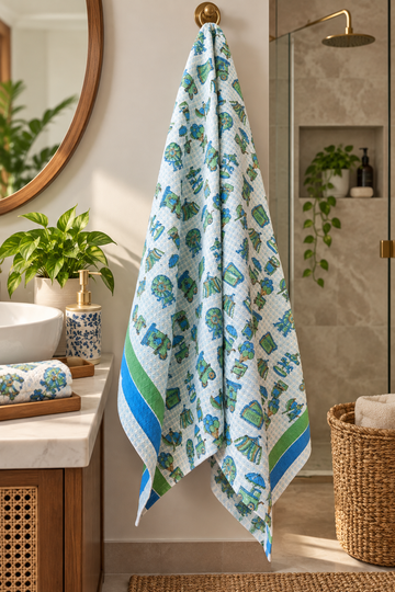 A patterned cotton bath towel hangs from a brass ring on a bathroom wall. The towel has a white base with a green and blue elephant print and striped borders.