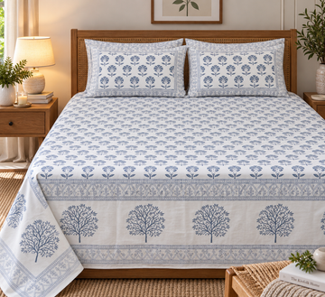A neatly made bed with a white bedsheet and matching pillowcases featuring a blue tree and floral block print pattern, styled in a warm, rustic bedroom setting with wooden furniture and bedside lamps.